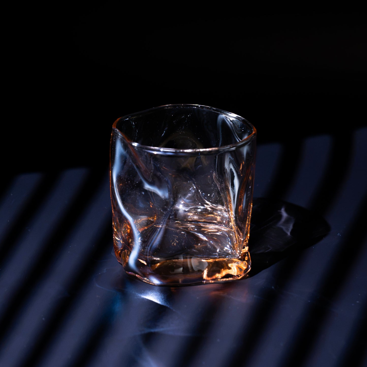 A clear textured whiskey glass with a faceted octagonal shape, containing amber liquid, set against a dark background with dramatic blue lighting and diagonal shadows.