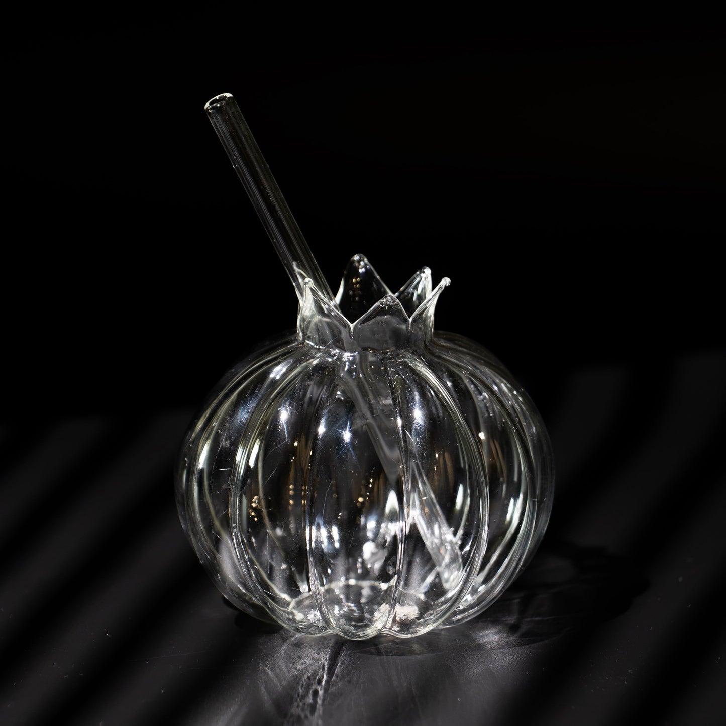 A clear ribbed glass bowl with a matching glass straw, photographed on a dark reflective surface against a black background.