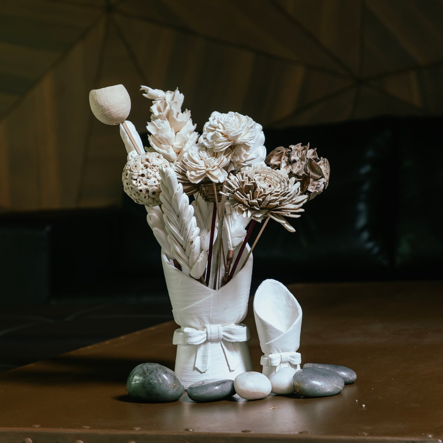 A decorative arrangement of dried flowers and leaves in a white ceramic vase, styled on a wooden table with smooth stones and a small matching white ceramic object beside it. The background is a dimly lit interior with dark furniture.