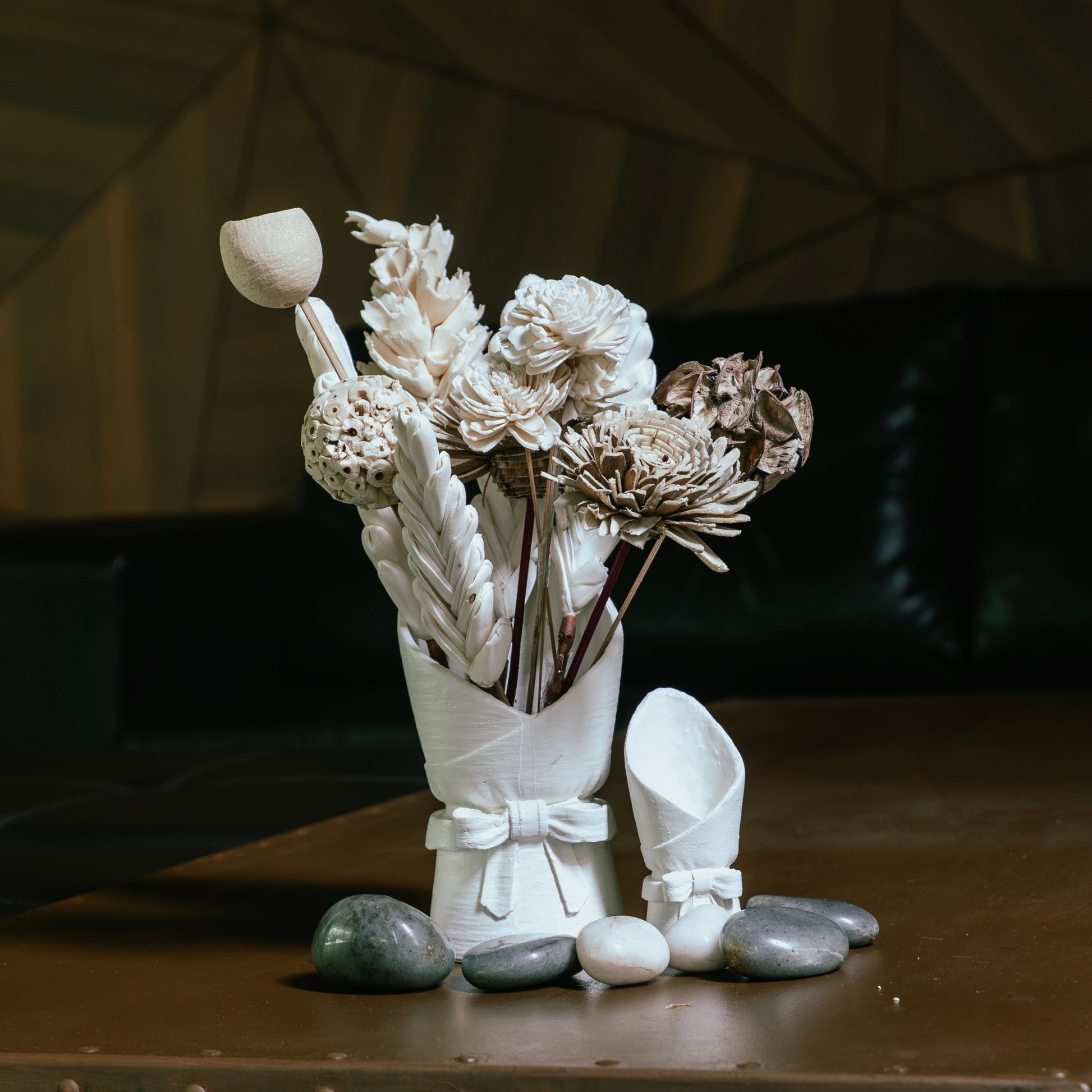 A decorative arrangement of dried flowers and leaves in a white ceramic vase, styled on a wooden table with smooth stones and a small matching white ceramic object beside it. The background is a dimly lit interior with dark furniture.