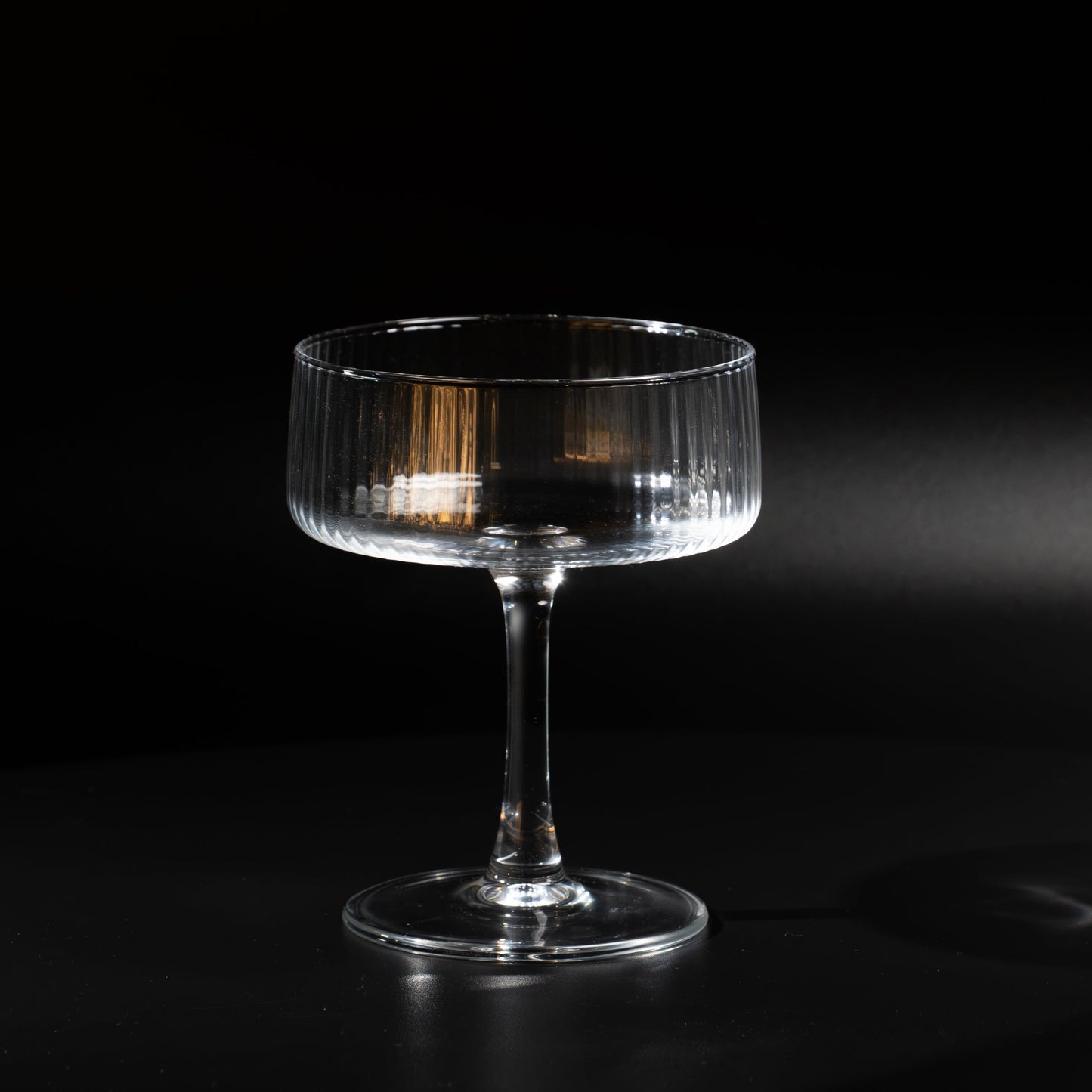 A clear ribbed-stripe coupe glass with a slender stem and circular base, photographed on a dark black background with dramatic side lighting.