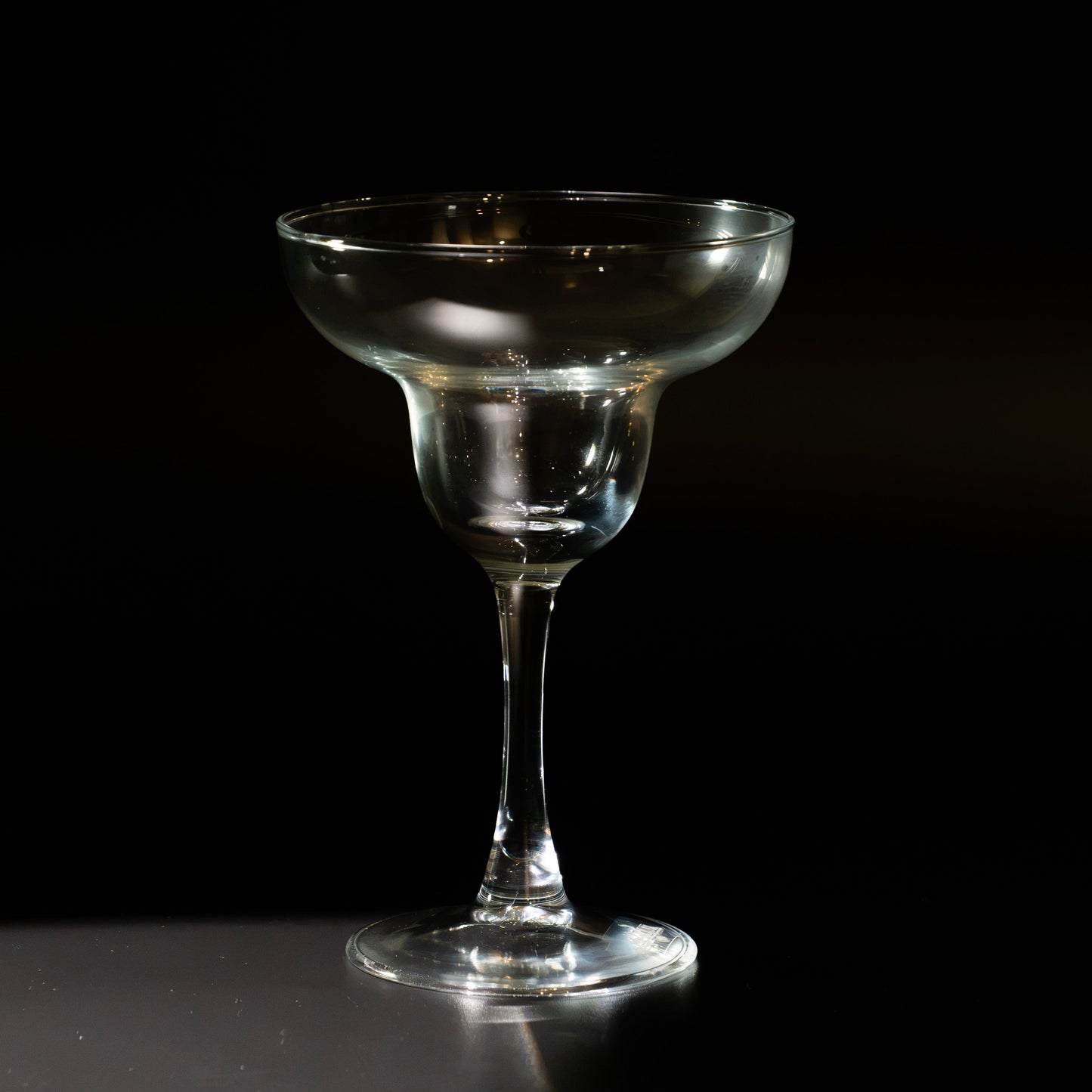A clear glass margarita coupe glass with a wide, shallow bowl and a long stem, standing on a dark reflective surface against a black background.