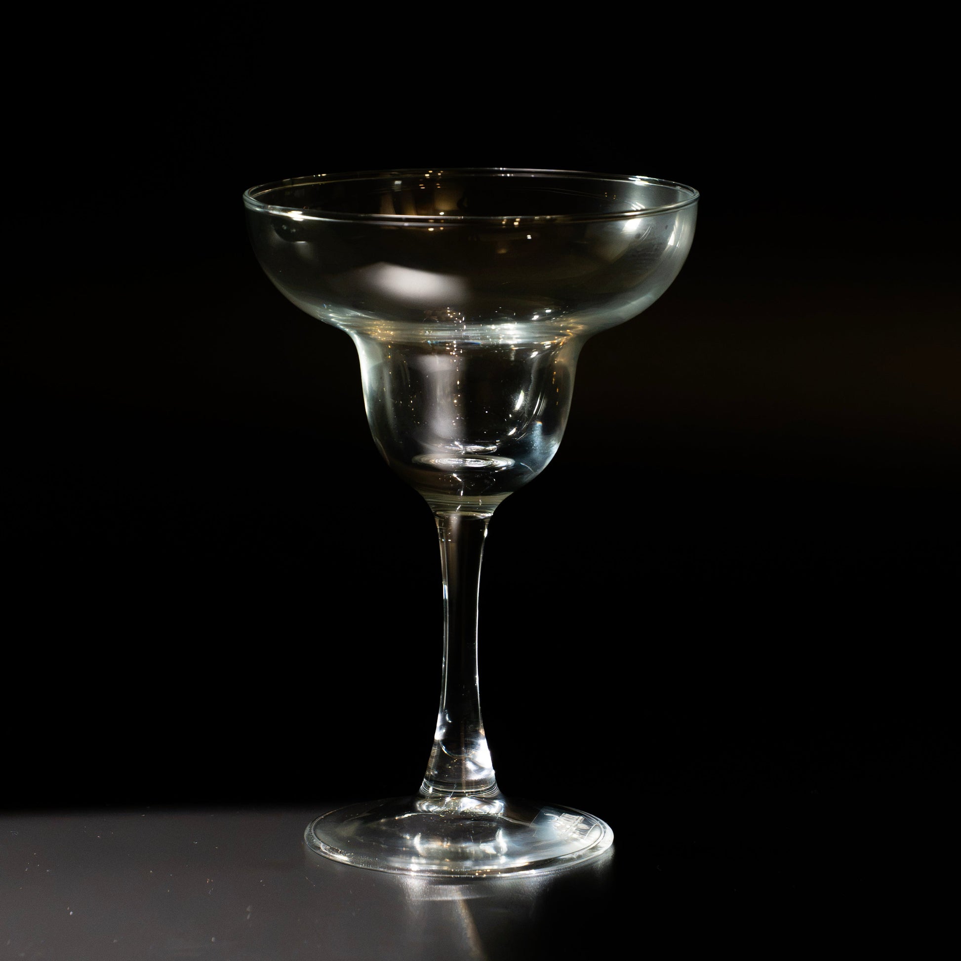 A clear glass margarita coupe glass with a wide, shallow bowl and a long stem, standing on a dark reflective surface against a black background.