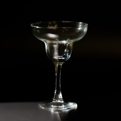 A clear glass margarita coupe glass with a wide, shallow bowl and a long stem, standing on a dark reflective surface against a black background.