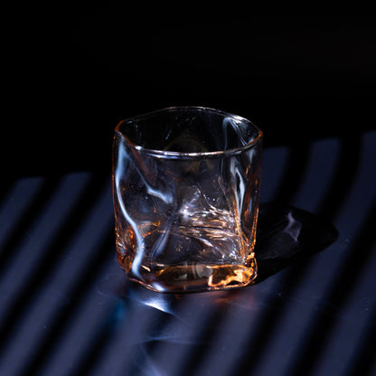 A clear textured whiskey glass with a faceted octagonal shape, containing amber liquid, set against a dark background with dramatic blue lighting and diagonal shadows.