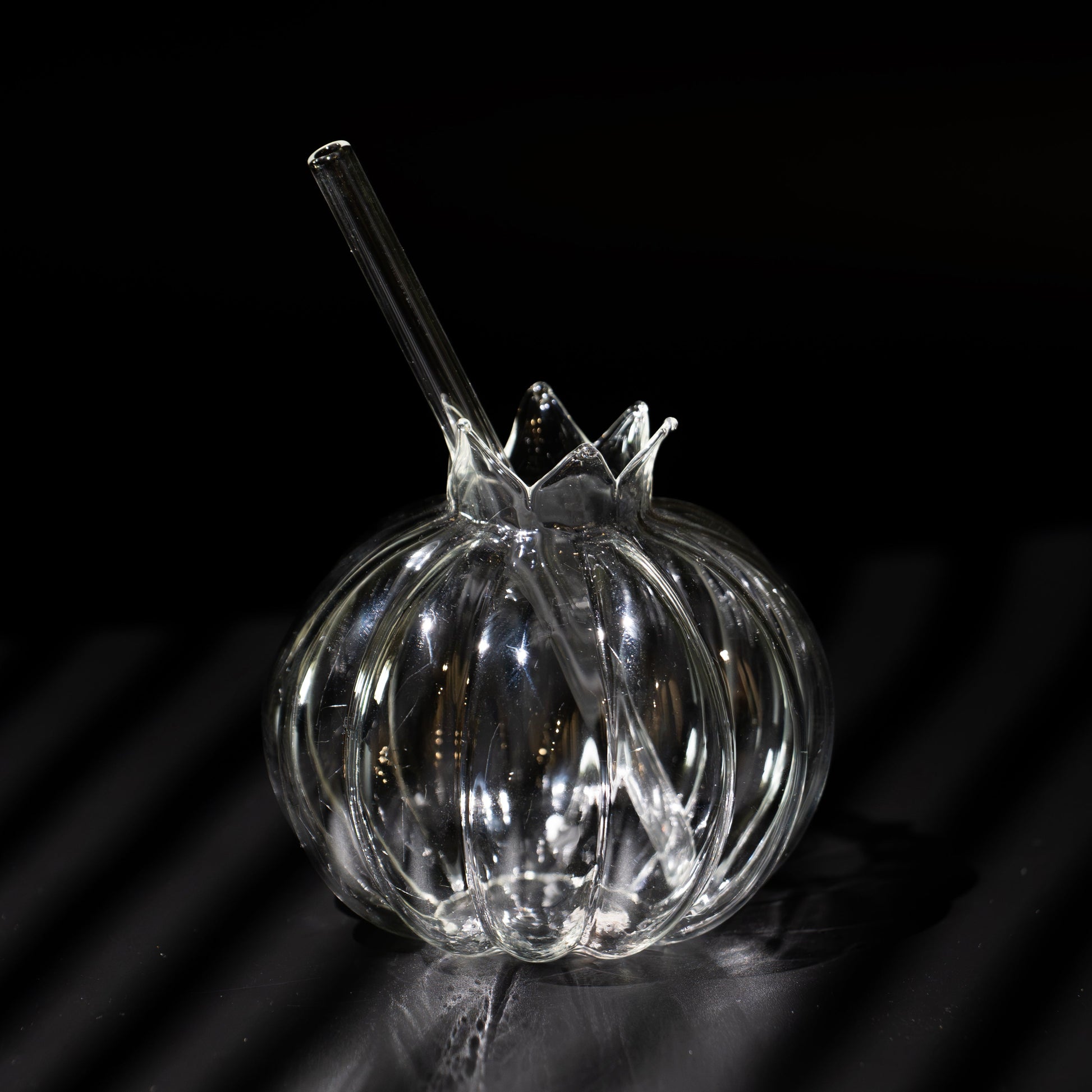 A clear ribbed glass bowl with a matching glass straw, photographed on a dark reflective surface against a black background.