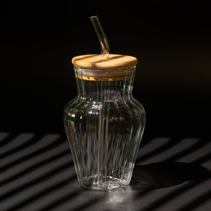 A ribbed clear glass bottle with a bamboo lid and a clear glass straw, set against a dark background with dramatic diagonal shadows.